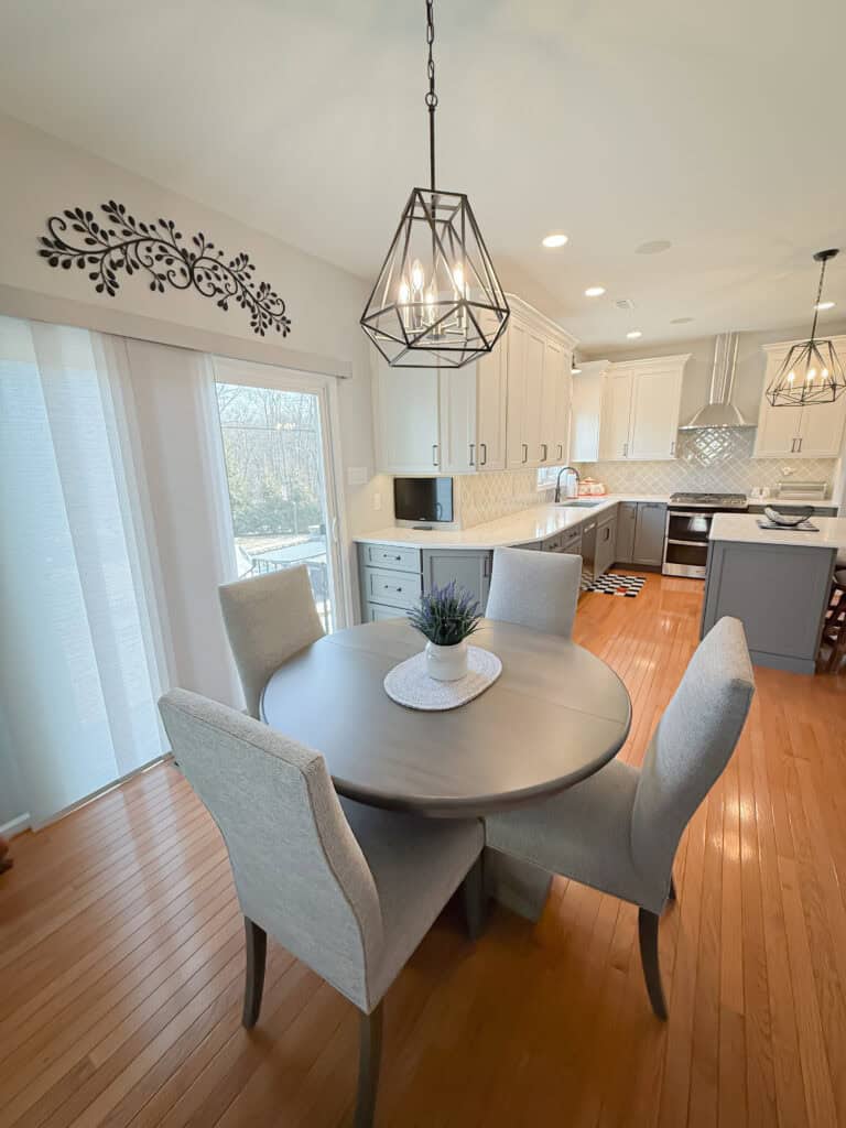 Modern kitchen and dining area with wooden floors, a round gray dining table with four upholstered chairs, pendant lights, white cabinets, and large sliding glass doors letting in natural light.