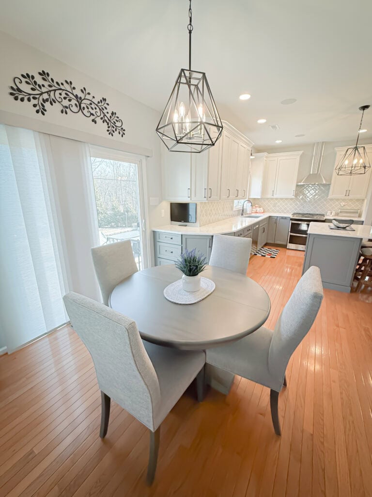 Bright, modern kitchen with white cabinets, light gray countertops, a round dining table with four upholstered chairs, hanging geometric light fixtures, and wood flooring. Sliding glass doors let in natural light.