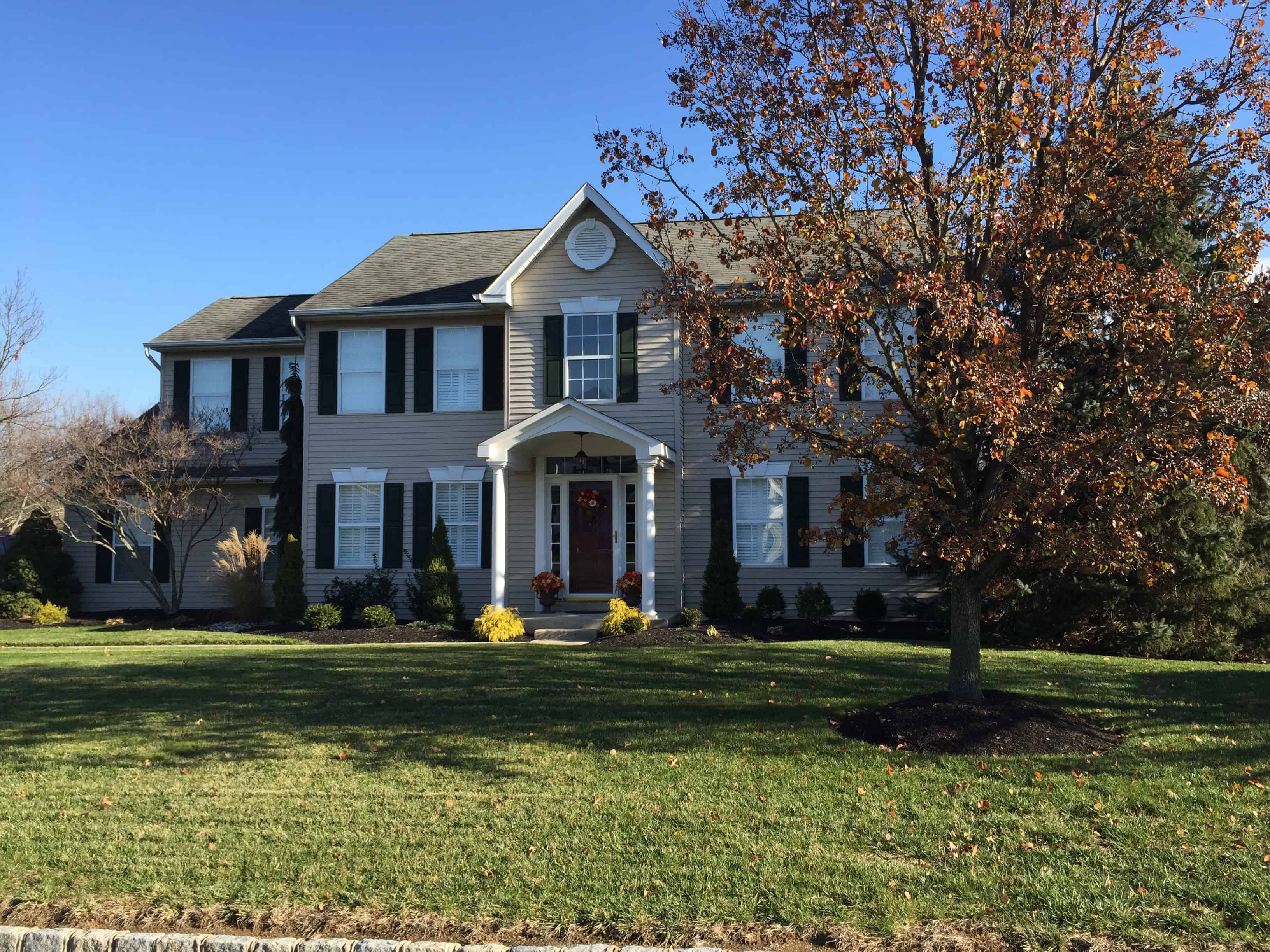Two-story suburban house with beige siding, dark shutters, and a red front door, surrounded by a well-kept lawn and a tree with autumn leaves under a clear blue sky.