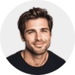 A young man with short brown hair and light stubble, wearing a dark shirt, is smiling at the camera against a plain light background.