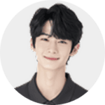 A young person with short, wavy black hair and fair skin smiles gently at the camera. They are wearing a dark collared shirt, and the background is plain and light-colored.