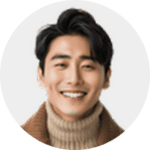 A young man with dark hair smiles at the camera. He is wearing a brown coat over a tan turtleneck sweater, and the background is light gray and out of focus.