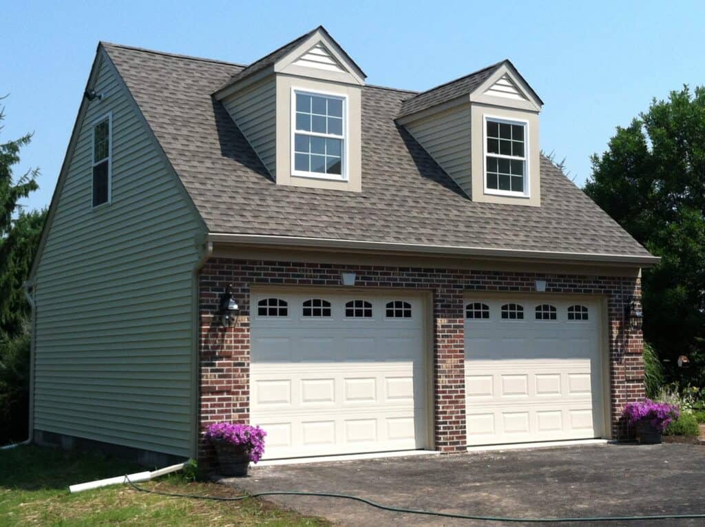 A two-car garage with a brick and beige siding exterior, featuring two white garage doors and two dormer windows above. Two planters with purple flowers sit on either side of the garage doors.