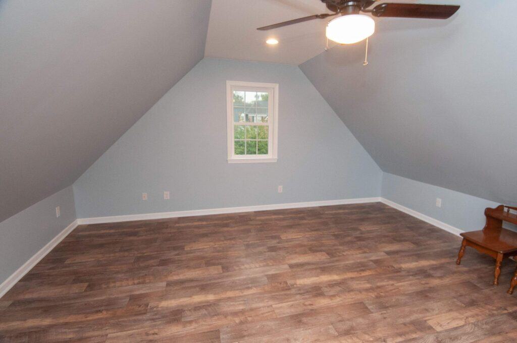 A spacious, empty attic room with slanted light blue walls, a ceiling fan with a light, a window, and dark wood flooring. There is a small wooden step stool in the corner.