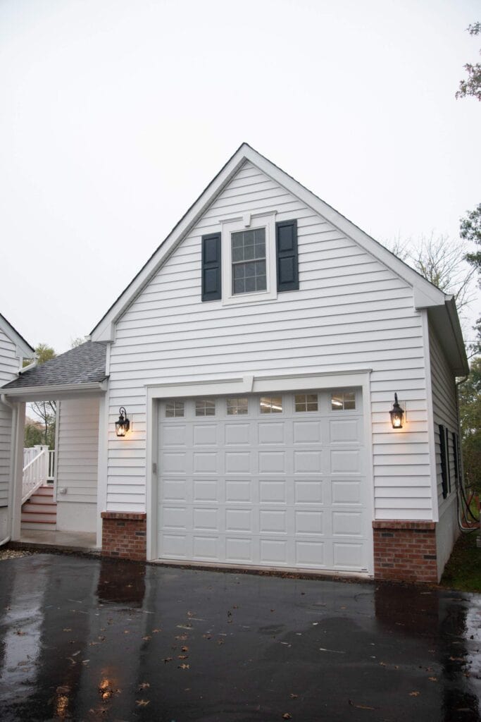 A white detached garage with an upper floor, featuring a large white garage door, an upstairs window with dark shutters, two wall-mounted lanterns, and a wet driveway reflecting light.