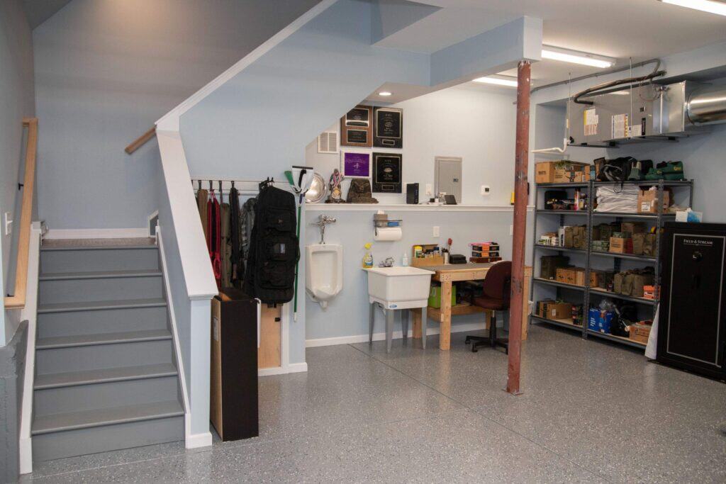 A tidy garage with gray stairs, a wall-mounted urinal and utility sink, storage shelves with boxes, a gun safe, a desk with a chair, and award plaques displayed on the wall.