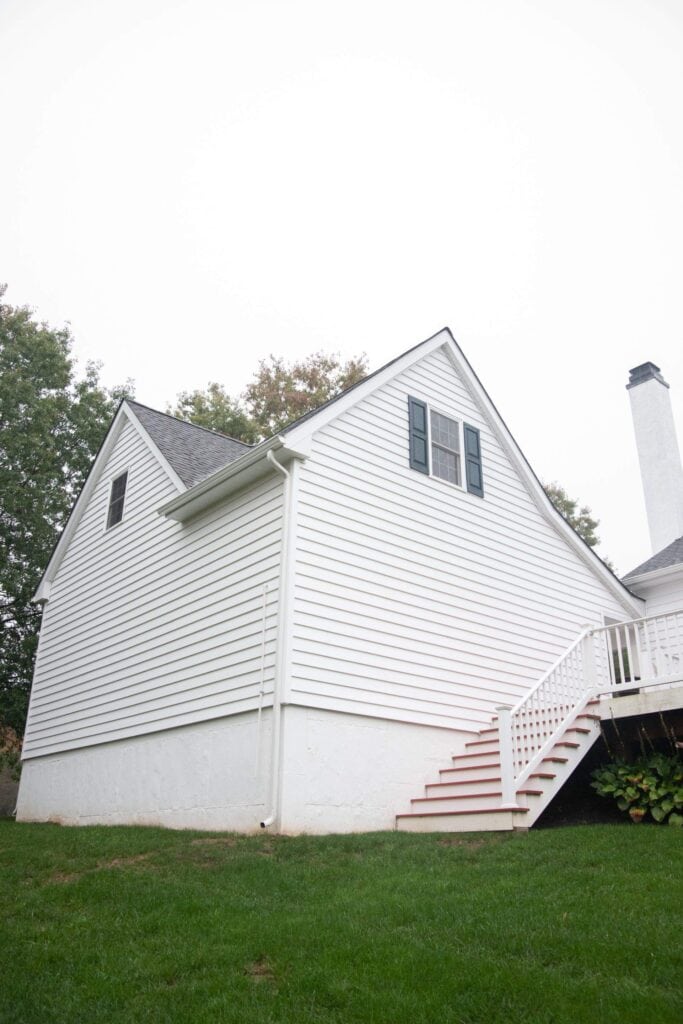 A white house with green shutters, a gable roof, and an exterior staircase leading to a small porch. The house is surrounded by green grass and trees, with an overcast sky above.