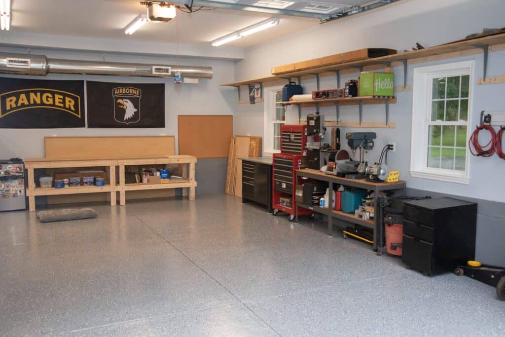 A clean, organized garage workshop with workbenches, toolboxes, shelves with tools and supplies, wall-mounted banners, and a window letting in natural light. The floor is spotless and gray.