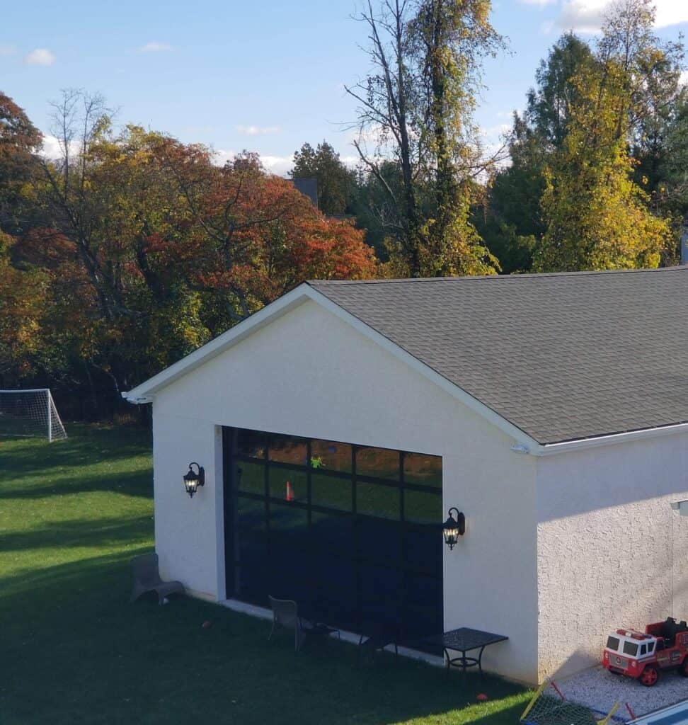 A small white building with a dark glass garage door sits on a grassy lawn, surrounded by trees with autumn foliage. Outdoor chairs and a table are near the wall, and children’s toys are visible on the right.