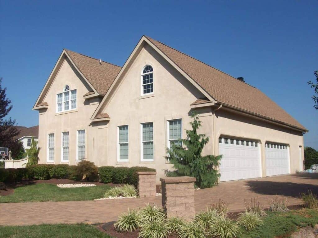 Two-story beige stucco house with a tall peaked roof, multiple white-trimmed windows, and a three-car garage. The front yard features green shrubs and a paved driveway under a clear blue sky.