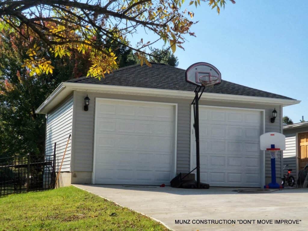 A double garage with white doors and gray siding sits under a clear sky. A basketball hoop stands on the driveway, with autumn leaves from a nearby tree visible at the top of the image.