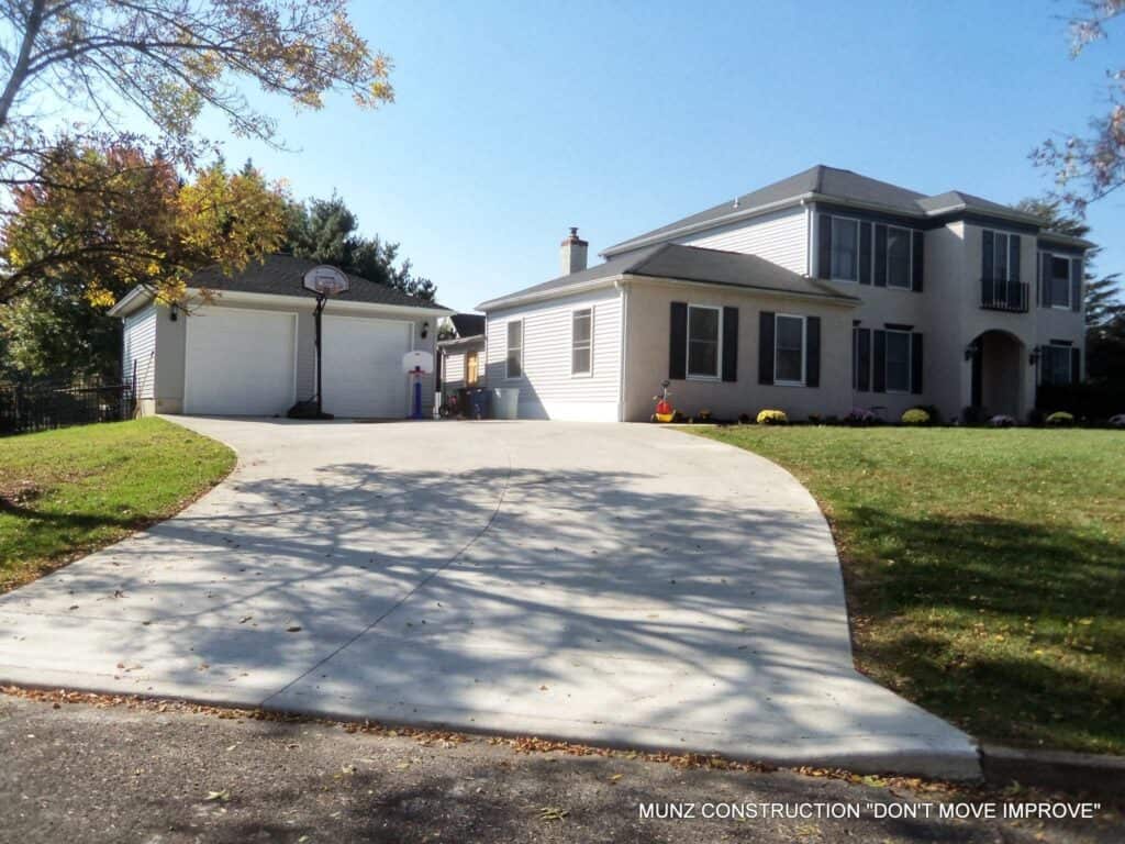 A two-story light-colored house with black shutters and a separate garage sits at the end of a wide concrete driveway, surrounded by grass and trees on a sunny day.