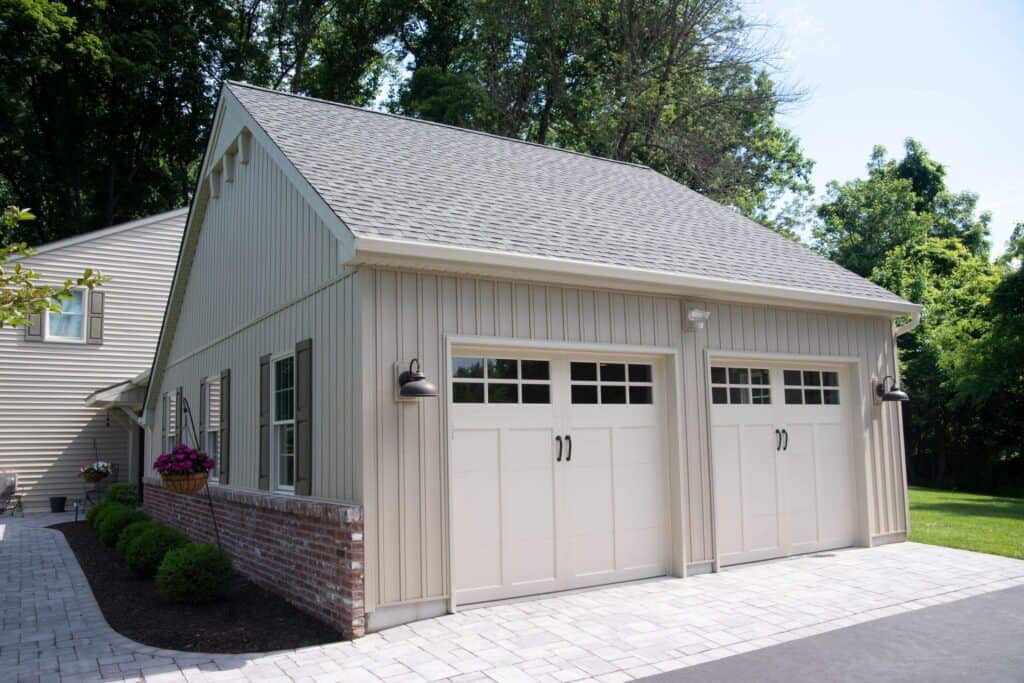 A beige detached garage with two white carriage-style doors, black lantern lights, and brick accents along the bottom, surrounded by landscaping and trees.