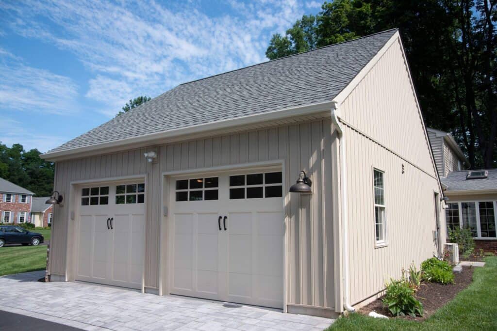 A beige detached two-car garage with white doors, exterior lights, a gabled roof, and paved driveway. The garage is surrounded by grass, small plants, and trees under a blue sky with clouds.