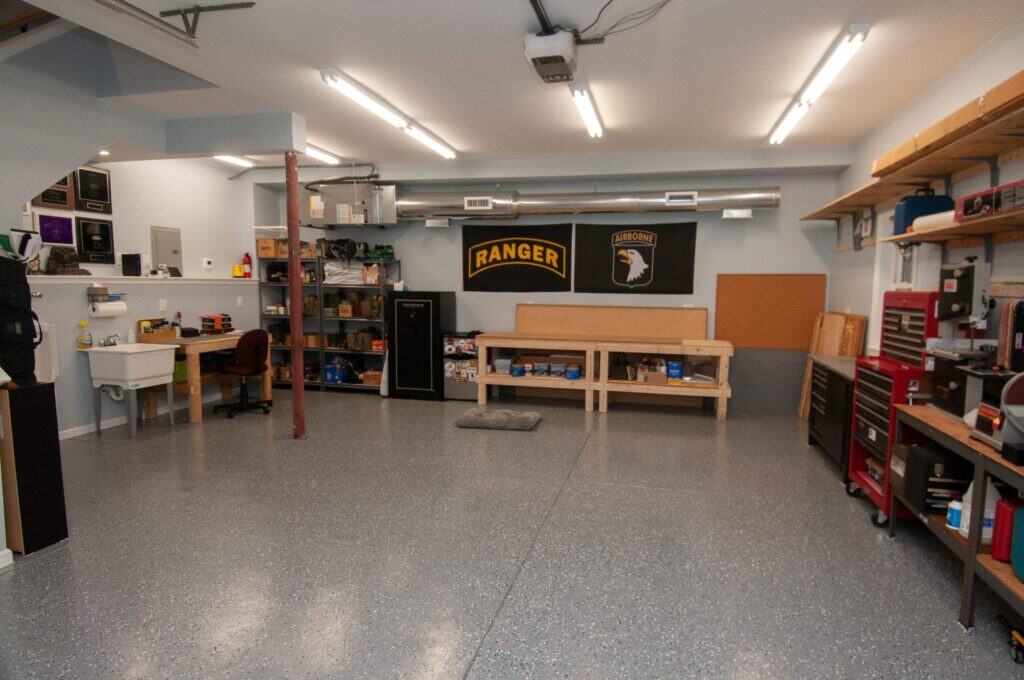 A clean, organized garage workshop with shelving, workbenches, a tool chest, a safe, a utility sink, and wall banners. The floor is speckled gray, and the space is brightly lit with overhead lights.