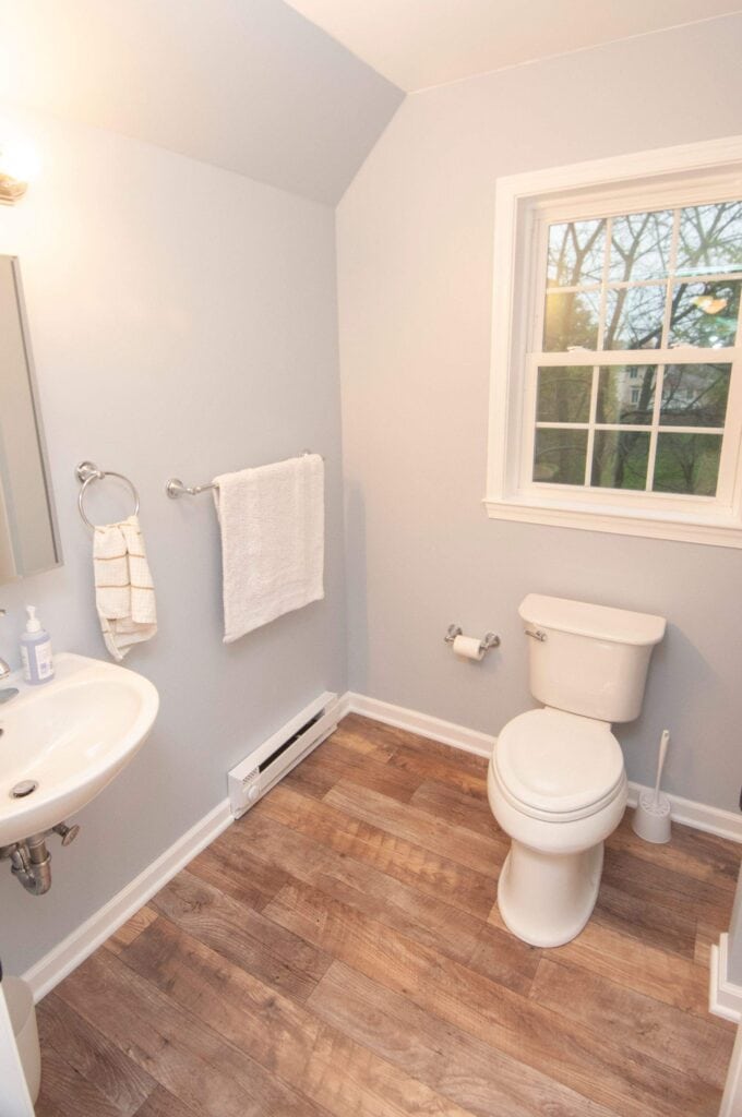 A small bathroom with light-colored walls, a white toilet, a wall-mounted sink, a towel ring with a towel, and a window letting in natural light. The floor has a wood-like finish.