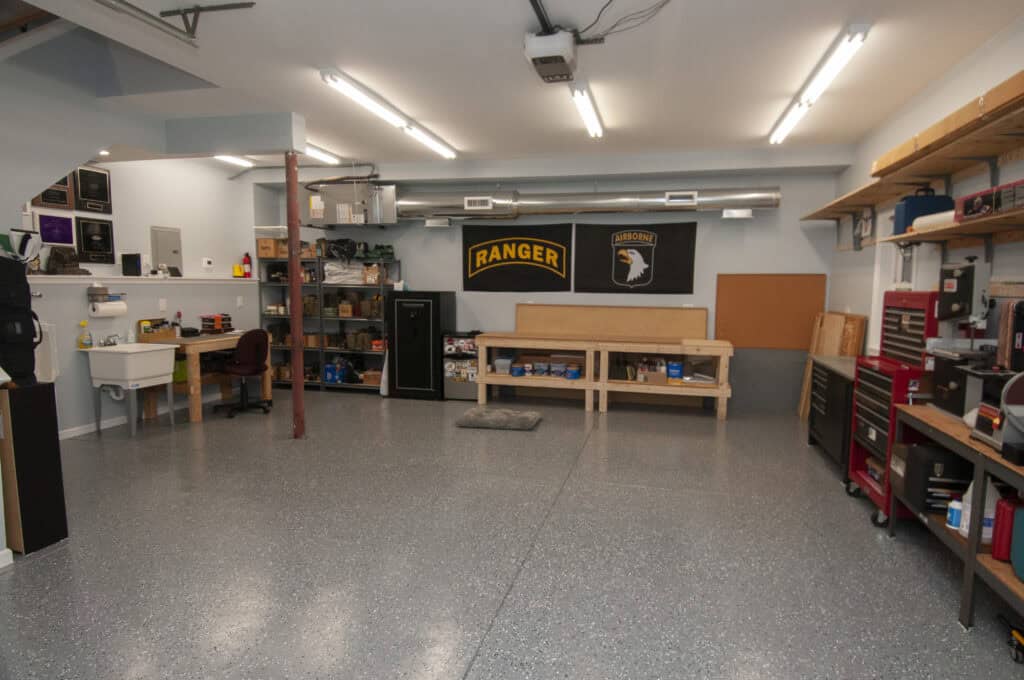 A clean, organized garage workspace with shelves, a workbench, storage cabinets, toolboxes, and banners on the wall. The floor is speckled gray, and fluorescent lights illuminate the space.