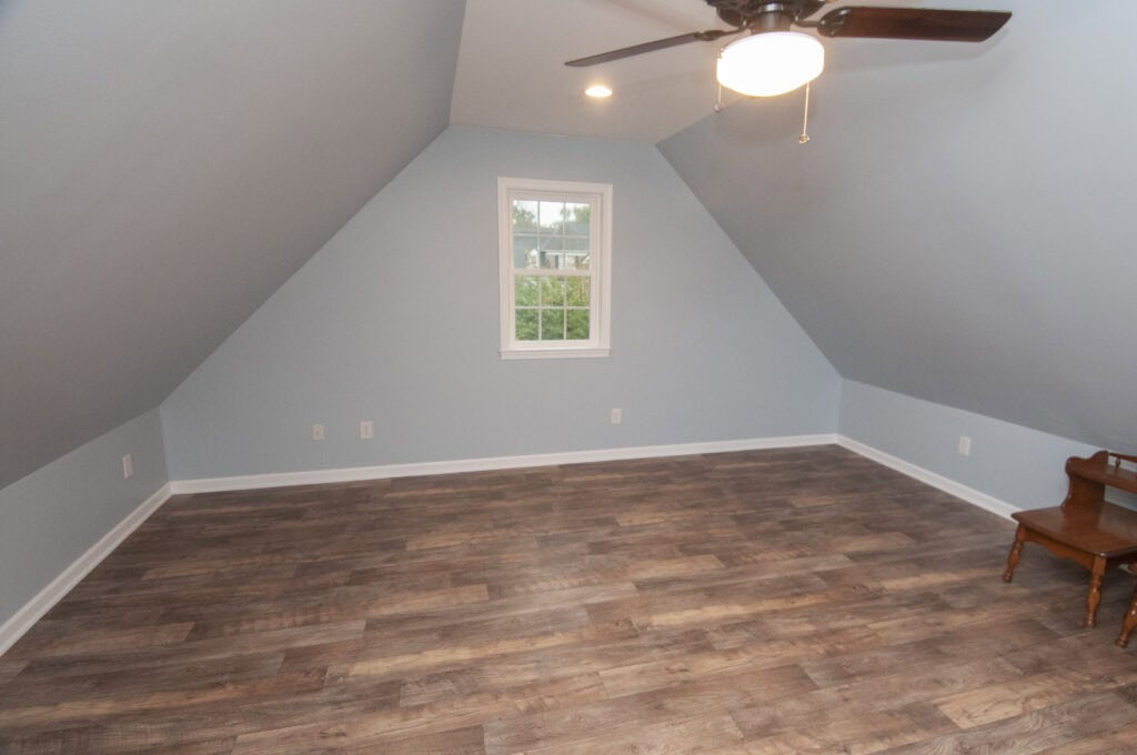 An empty attic room with light blue walls, a single window, wood-look flooring, a ceiling fan with a light, and a small wooden bench in the corner.