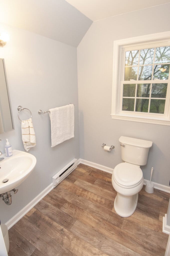 A small bathroom with light-colored walls, a white toilet, a wall-mounted sink, a towel ring with towels, a toilet paper holder, a window, and wood-look flooring.