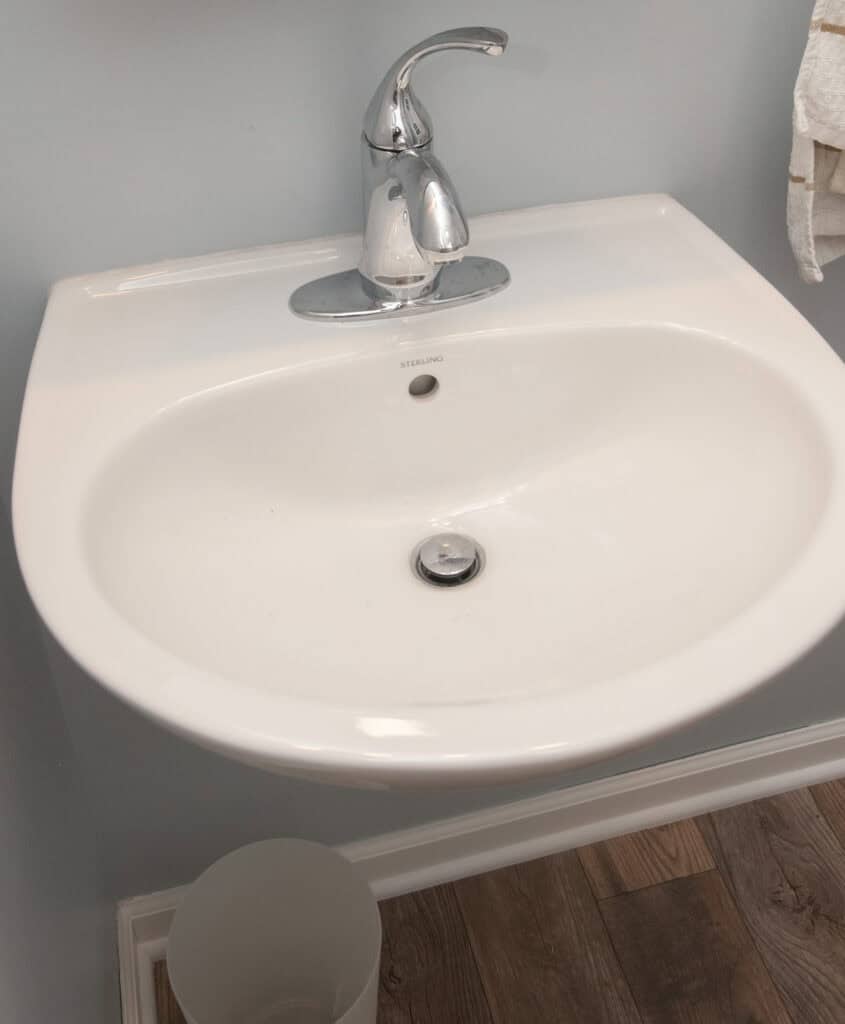 A white ceramic bathroom sink with a silver faucet, set against a light blue wall. A white towel hangs to the right, and the sink is above wood-patterned flooring with a small white trash can below.