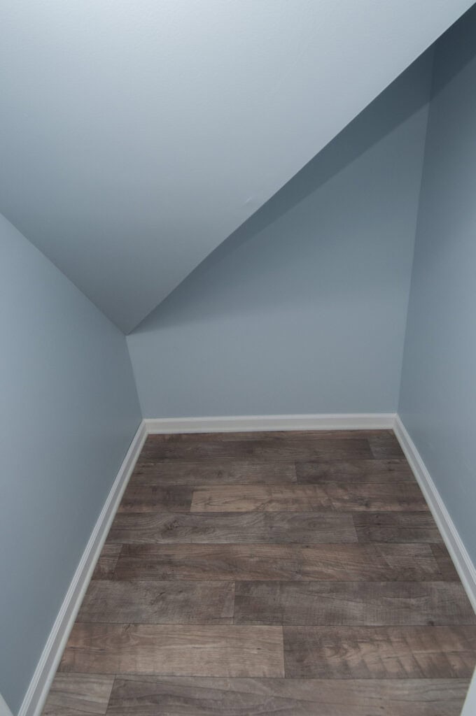 A small empty room with blue walls, a white baseboard, and slanted ceiling, featuring wood-patterned flooring.