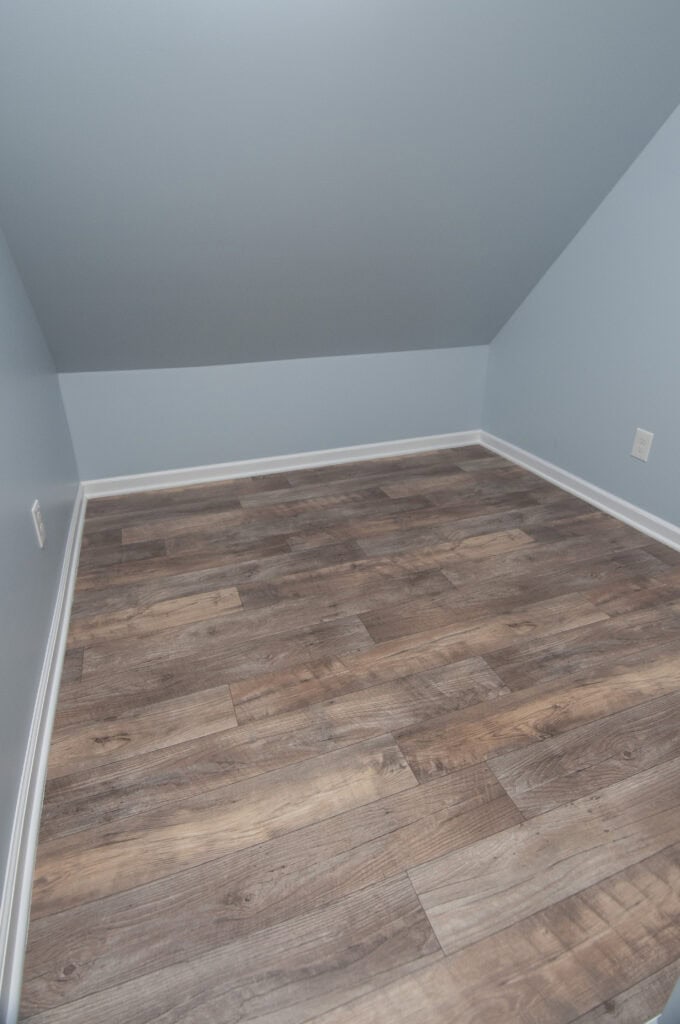 A small, empty room with light blue walls, white baseboards, and wood-look vinyl flooring. The ceiling slopes down on one side, creating a cozy, attic-like space.