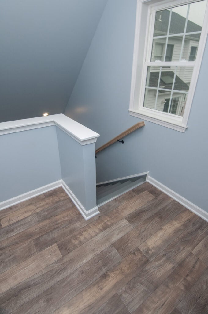 A staircase with a wooden handrail descends from a landing with light brown wood flooring and blue walls. A window shows neighboring houses outside. The area is clean and well-lit.