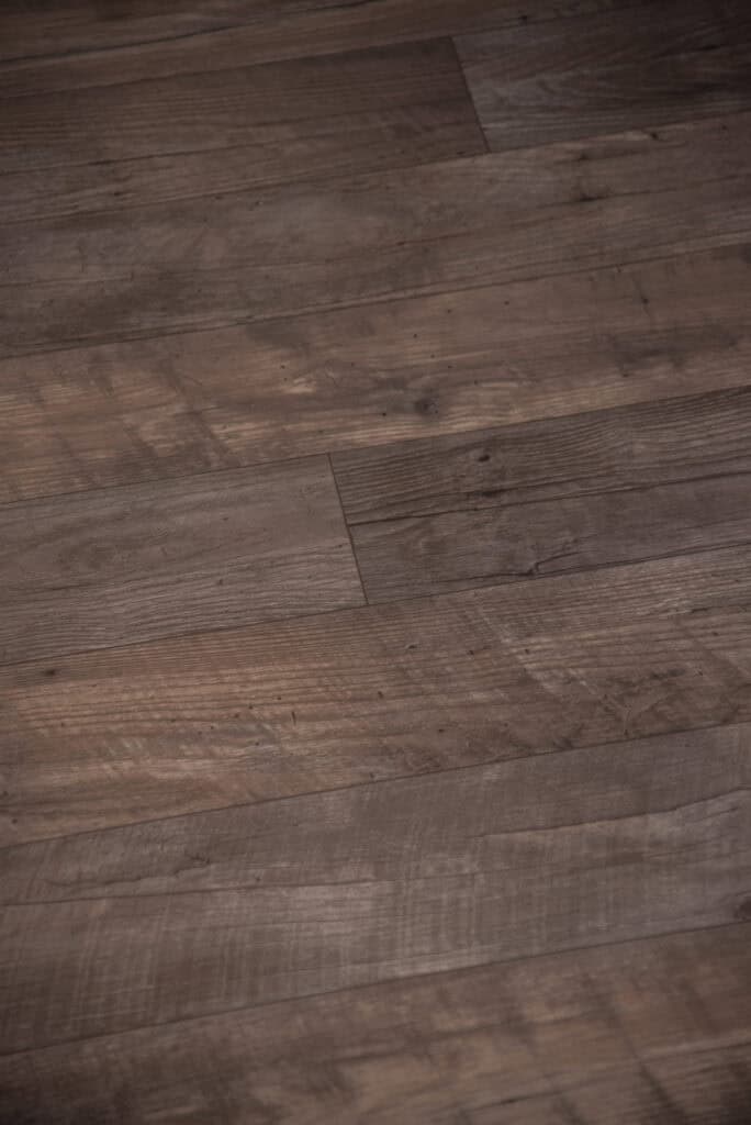 Close-up view of a wooden floor with medium brown planks, showing natural grain patterns, knots, and some subtle scuff marks. The planks are laid lengthwise with varying shades of brown.
