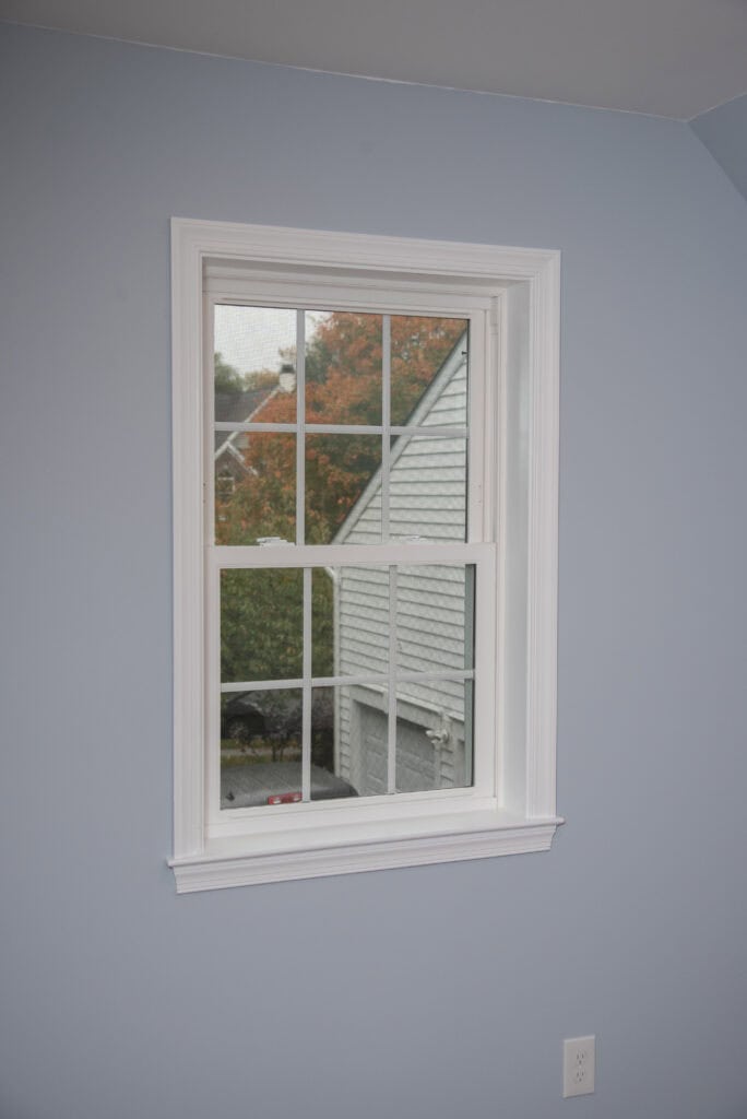 A white-framed window in a light blue wall shows the side of a gray house and orange autumn foliage outside. An electrical outlet is visible below the window.