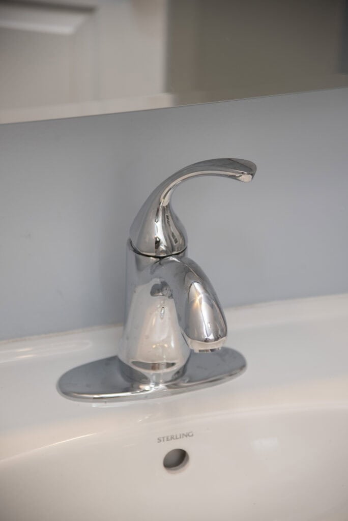 A close-up of a modern, chrome bathroom faucet mounted on a white sink with a light gray wall and a mirror in the background.