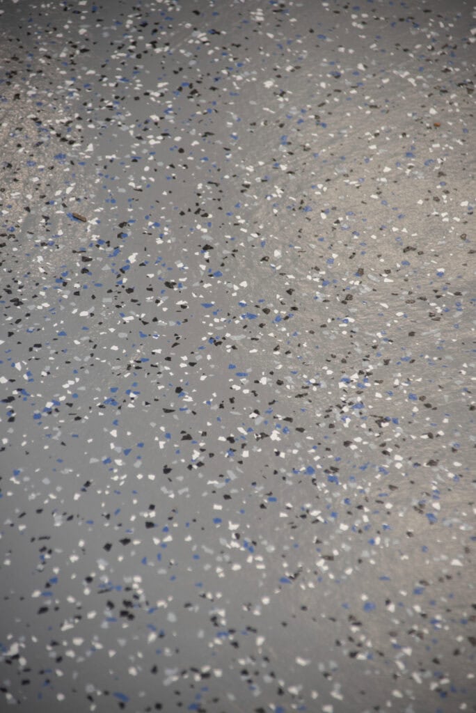 Gray concrete floor with a speckled pattern of scattered black, white, gray, and blue chips, resembling an epoxy or decorative flake coating, creating a textured and colorful surface.