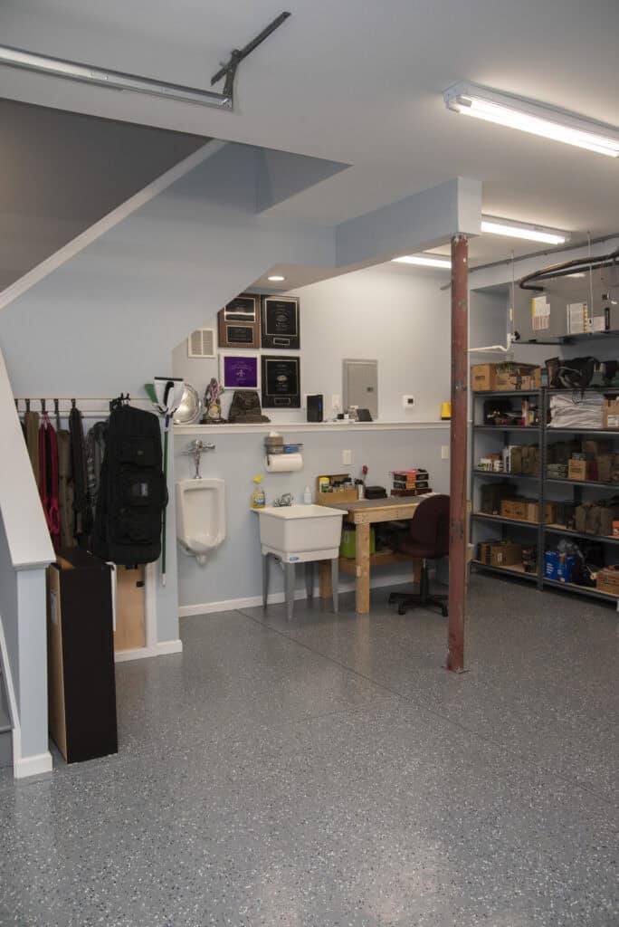 A clean, organized garage with a utility sink, urinal, desk, shelves with various items, wall plaques, and coats hanging under a staircase. The concrete floor is speckled gray.