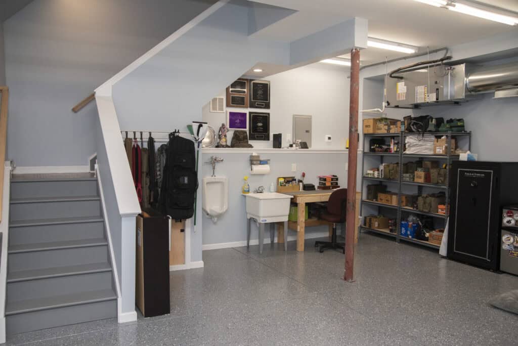 A clean, organized garage with gray walls and floor, featuring a staircase, utility sink, urinal, work desk, wall-mounted plaques, shelves with tools and supplies, and a black safe.