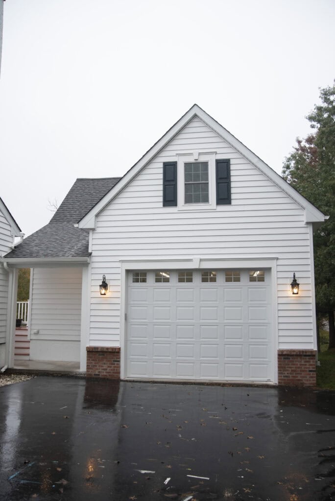 A white house with a single-car garage, black shutters, brick trim, and outdoor wall lanterns. The driveway is wet, and trees are visible in the background on a cloudy day.