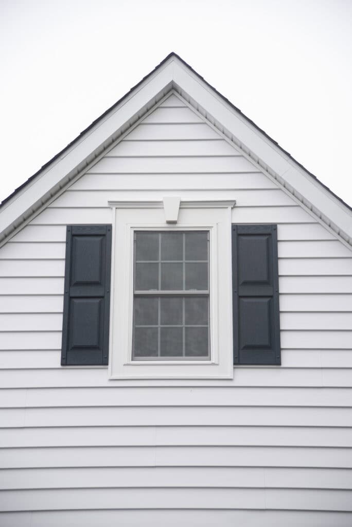 The image shows the upper portion of a white house with horizontal siding, a single window with black shutters on each side, and a triangular roof peak above.