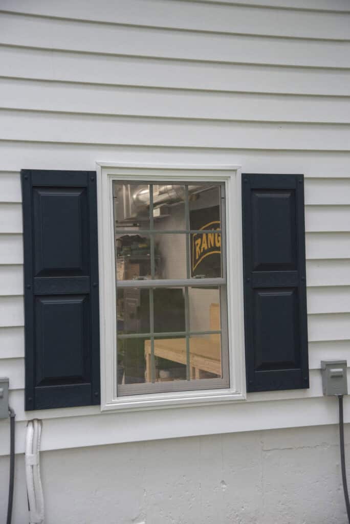 A window with dark shutters set in white siding, reflecting an indoor workshop with shelves, pipes, and a yellow RANGER sign visible through the glass.