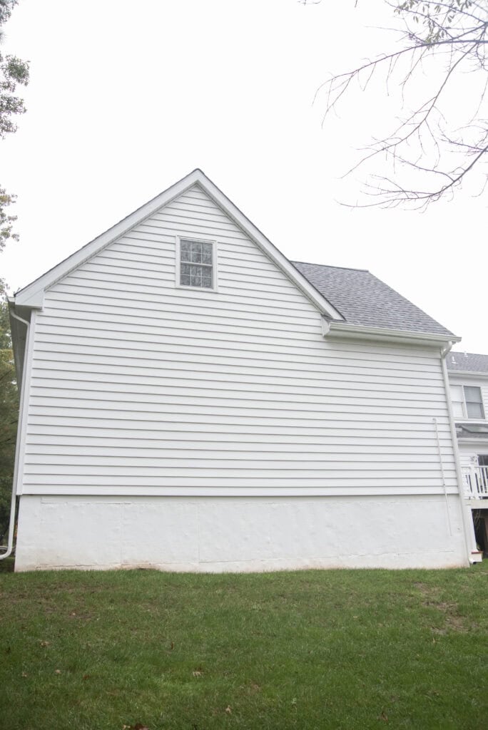 The image shows the side of a white house with horizontal siding, a single small window near the roof peak, and a grassy lawn in the foreground. The sky appears overcast.