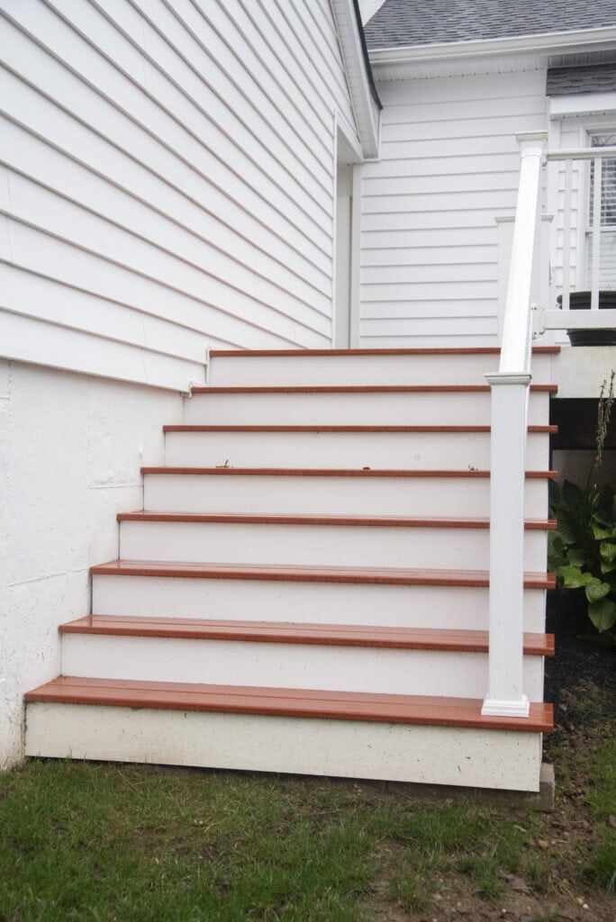 A set of outdoor wooden steps with white risers and reddish-brown treads leads up to a white house, with a single white handrail on the right side.