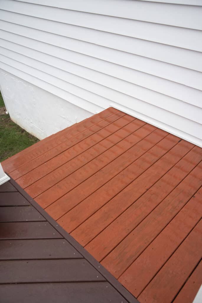 A clean, newly stained wooden deck in two colors—brown and reddish-orange—adjoins the white vinyl siding of a house. The deck appears wet from recent rain. Grass is visible next to the base of the house.