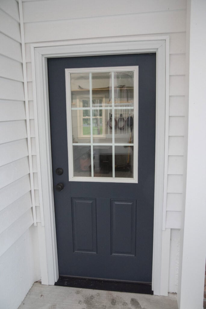 A dark blue exterior door with a windowpane of six glass panels, set in a white frame, on a porch with white siding. Some items can be seen through the window inside the house.