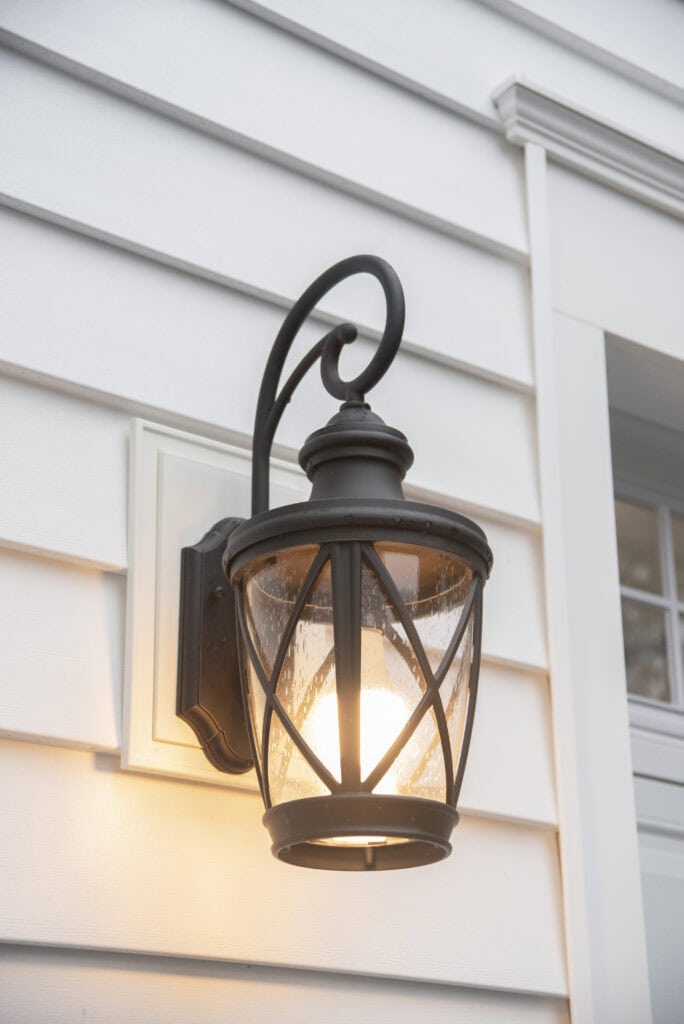 A black metal outdoor wall lantern with a glass shade is mounted on white horizontal siding, illuminating the exterior of a house near a window.