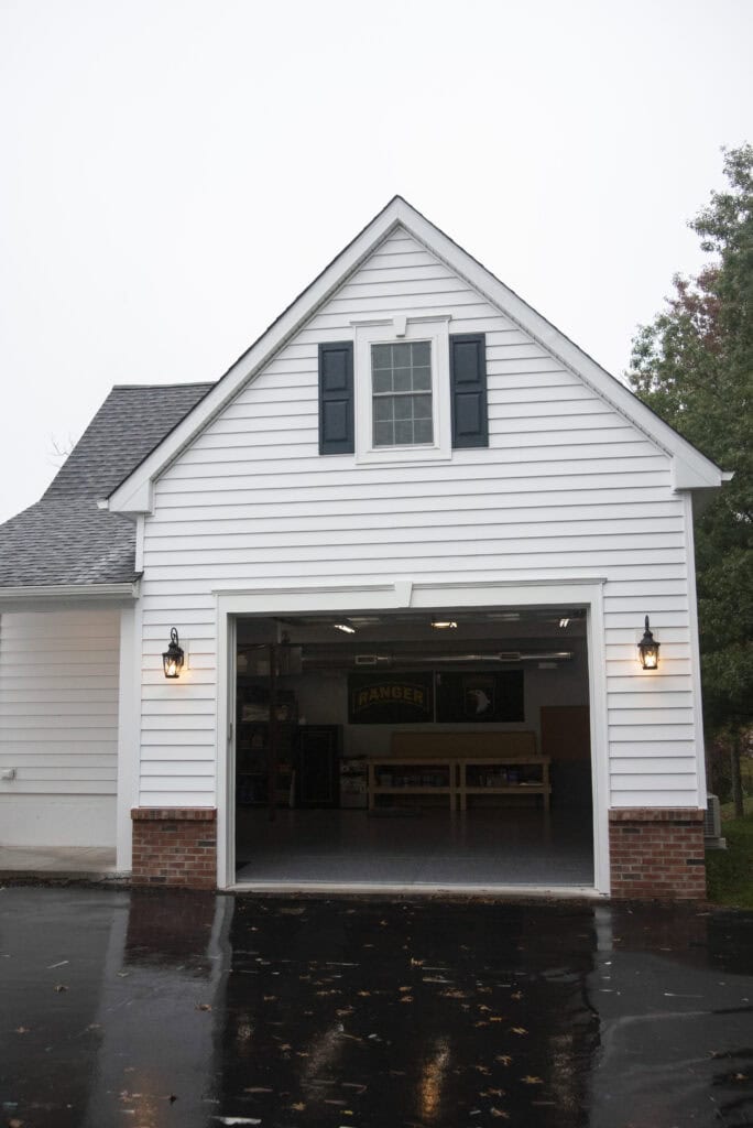 A white garage with a peaked roof and a single window above the open garage door, revealing a clean and organized interior. Two lantern-style lights are mounted on either side of the entrance.