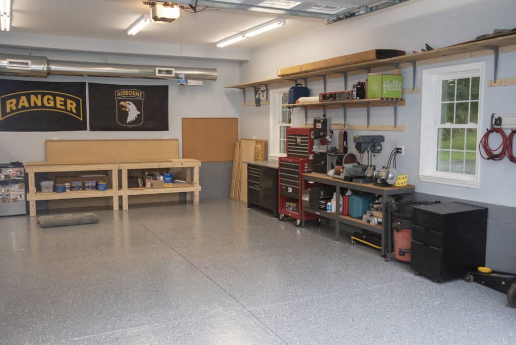 A clean, organized garage workshop with tools, toolboxes, and shelves along the walls, two military banners, a workbench, and a window letting in natural light.