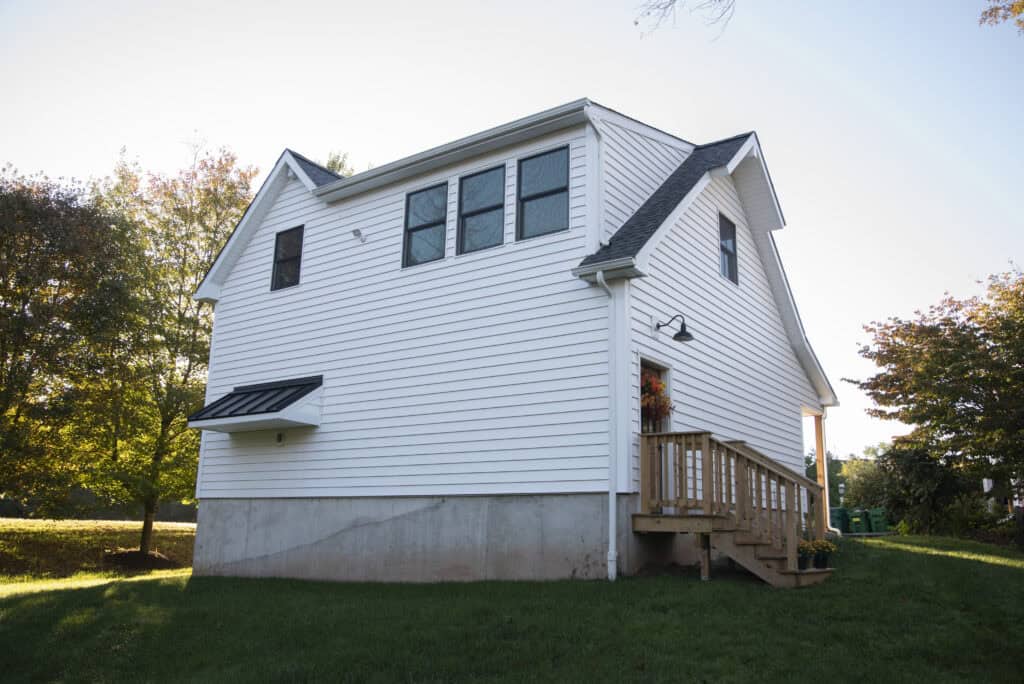 A white two-story house with black roof shingles, elevated on a concrete foundation, sits on a grassy lawn. Wooden steps lead to the front door, and autumn trees are visible in the background under clear skies.