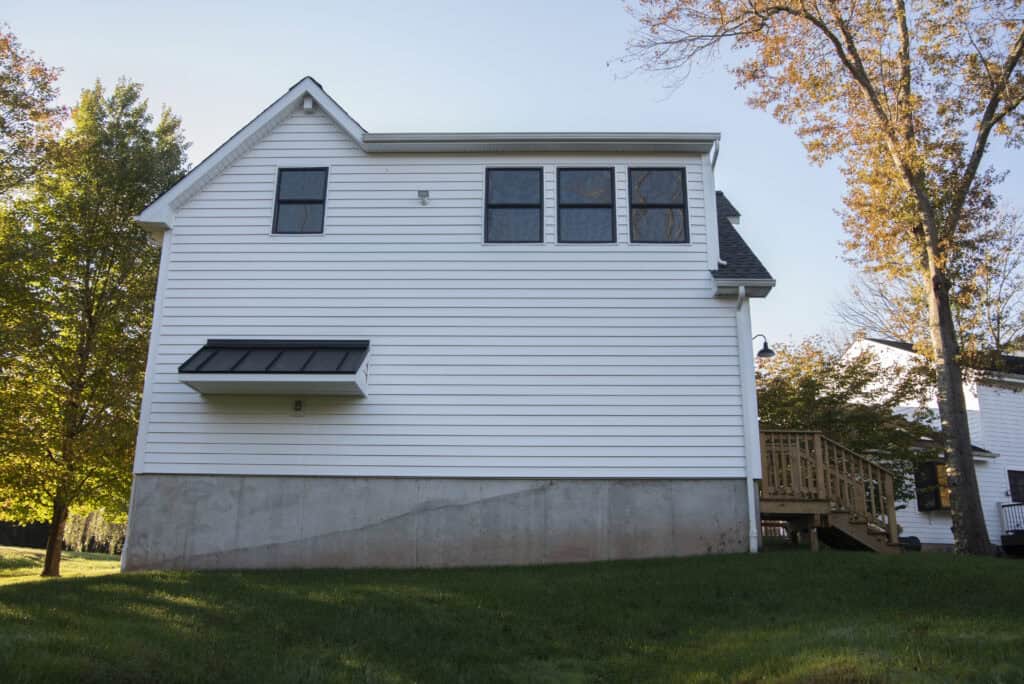 A white house with black trim and several windows, elevated on a concrete foundation, sits on a grassy lawn with trees in the background. A wooden staircase leads to a side entrance.