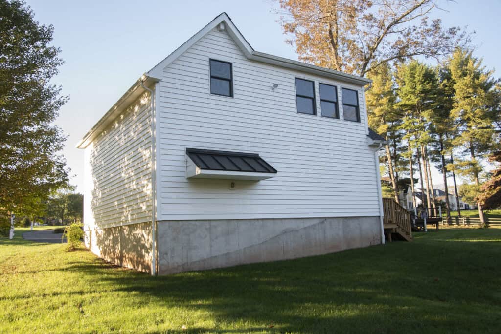 A white house with a concrete foundation and three windows, one with a small black awning, featuring a grassy lawn and trees in the background on a sunny day.