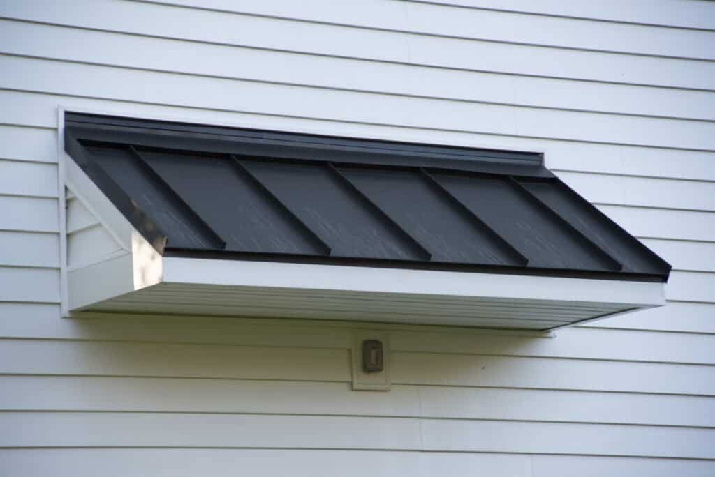 A black metal awning with a white frame is attached to a white vinyl-sided building. There is a small rectangular light fixture mounted beneath the awning.