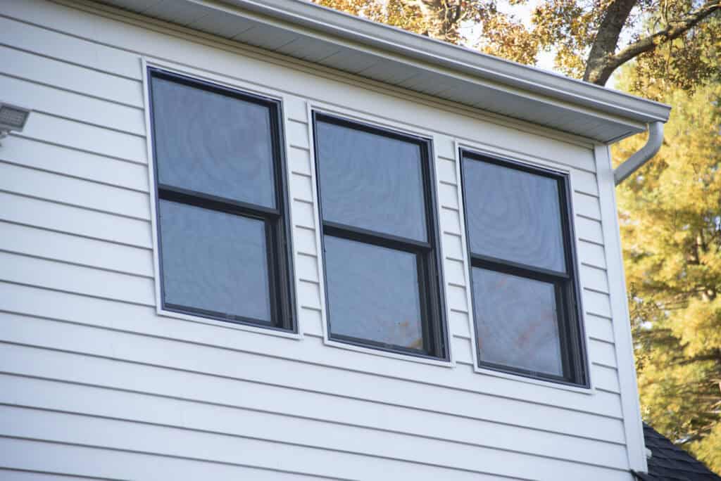The image shows three large rectangular windows with black frames on the second story of a white house with horizontal siding, under a white roofline. Trees with autumn leaves are visible in the background.