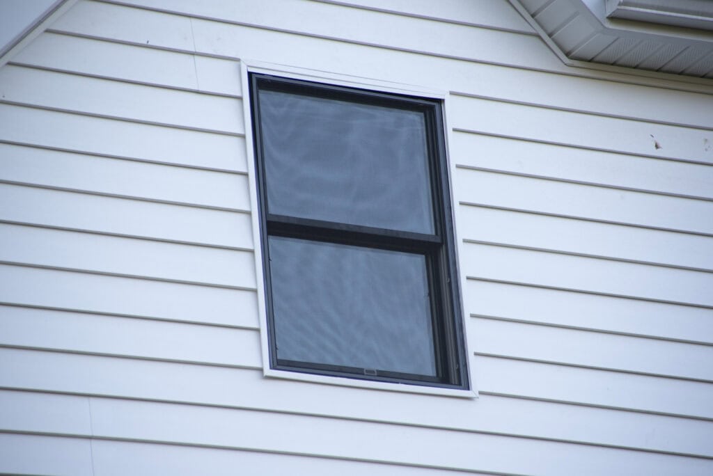 A double-hung window with dark trim is set into the white siding of a house. The window is closed and reflects the sky, while the white siding panels run horizontally across the wall.
