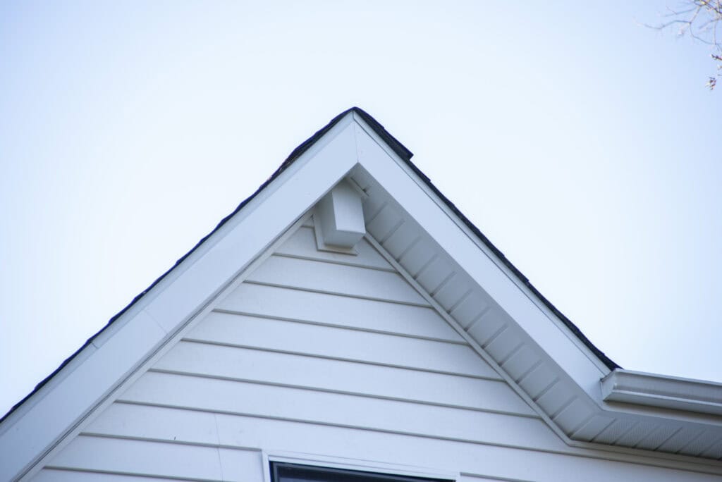 A close-up of the peak of a white house’s gable roof, showing white siding, dark roof shingles, and a small rectangular attic vent against a clear sky.